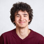 Photo of handsome unshaven guy looks with pleasent expression directly at camera, wears fashionable maroon t shirt, stands against white studio wall. People, lifestyle and fashion concept. Copy space.